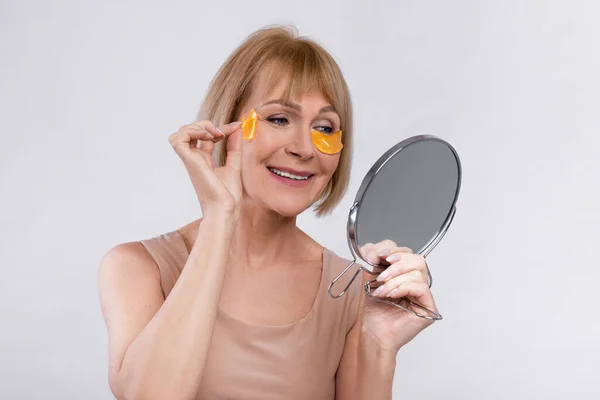 Smiling mature lady using golden hydrogel eye patches near mirror over light studio backgroud. Beauty and skincare — Stock Photo, Image