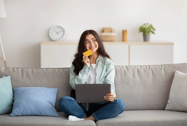 Mulher indiana com laptop pc e cartão de crédito pensando sobre suas compras on-line, sentado no sofá em casa, comprimento total — Fotografia de Stock