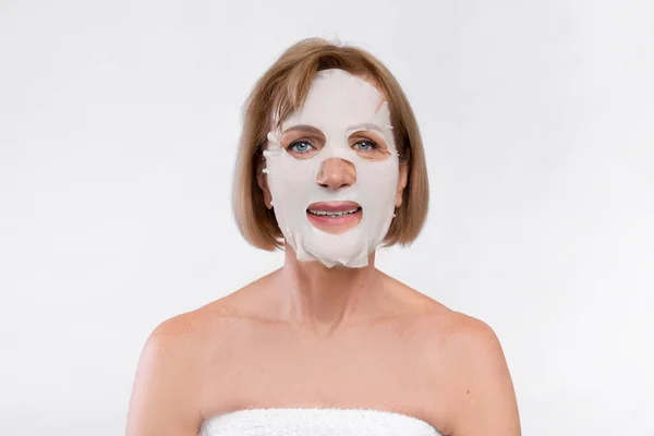 Portrait of mature Caucasian woman wearing cotton facial mask on light studio background. Domestic beauty treatment — Stock Photo, Image