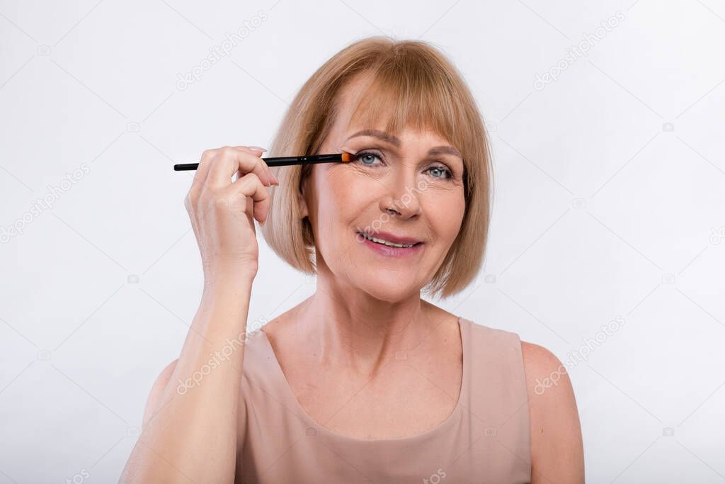Beauty portrait of cheerful senior woman applying eye shadow with brush over light studio background