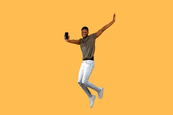 Vreugdevolle Afrikaanse amerikaanse man springen over gele achtergrond en toont smartphone met zwart scherm naar camera. — Stockfoto