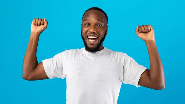 Vreugdevolle Afrikaanse man schudden vuisten kijken naar camera, blauwe achtergrond — Stockfoto