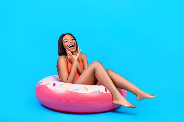 Portrait of excited black lady in bikini touching her face, sitting in donut shaped inflatable ring on blue background