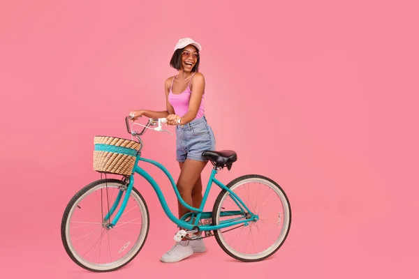 Charming young black lady in summer wear standing with classic bicycle and looking at empty space, pink background — Stock Photo, Image