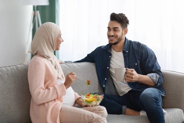 Mulher Islâmica grávida comendo salada de legumes enquanto descansa no sofá com o marido — Fotografia de Stock