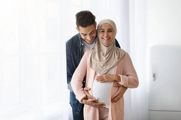 Amar marido árabe abrazando a su esposa musulmana embarazada mientras está de pie cerca de la ventana — Foto de Stock