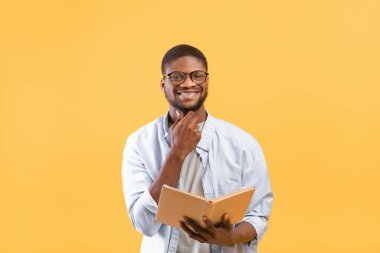 Gözlüklü mutlu siyah öğrenci okuma kitabı açıyor, sarı arka plan sınavına çalışıyor.
