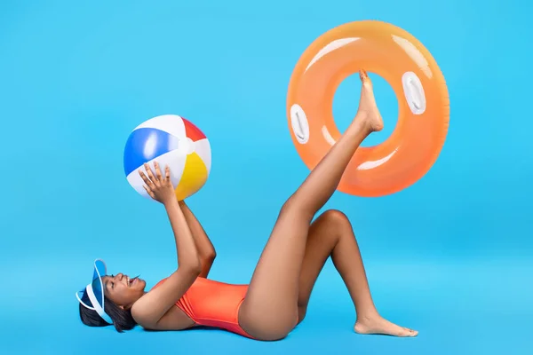 Full length shot of joyful black lady in swimwear playing with inflatable ring and beach ball on blue studio background — Stock Photo, Image