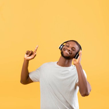 Havalı Afrikalı Amerikalı adam kablosuz kulaklıklarıyla müzik listesinin keyfini çıkarıyor ve sarı arka planda dans ediyor.