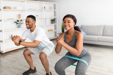Genç, formda, lastik bantlı, evde çömelme hareketleri yapan siyah çift eğitimi alıyor.