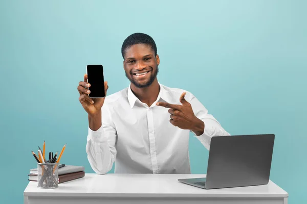 Afrykański freelancer siedzi w miejscu pracy z laptopem i wskazuje na komórkę z pustym ekranem, niebieskie tło — Zdjęcie stockowe