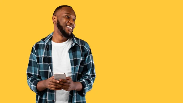 Happy Black Male Posing With Smartphone Looking Aside, Yellow Background