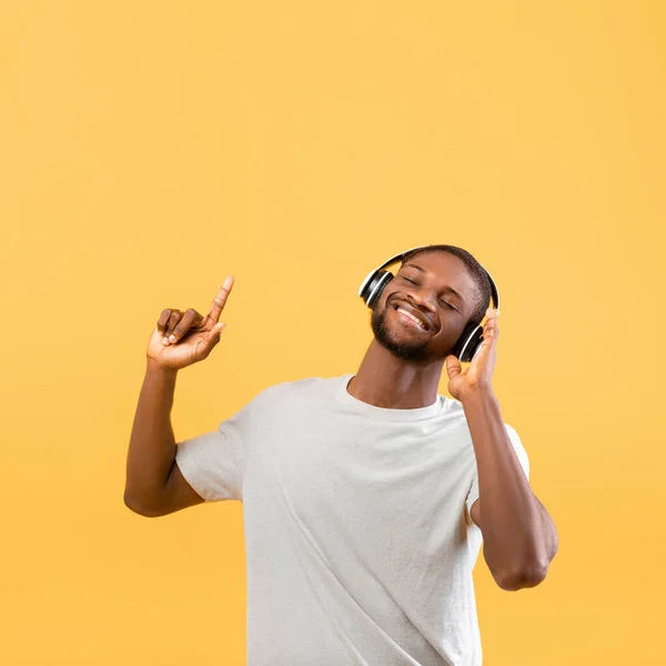 Cool homem americano africano desfrutando de sua lista de reprodução em fones de ouvido sem fio e dançando sobre fundo amarelo, espaço livre — Fotografia de Stock