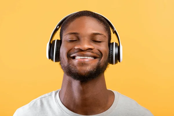 Fan de la música. Hombre afroamericano pacífico en auriculares escuchando música con los ojos cerrados, de pie sobre fondo amarillo — Foto de Stock
