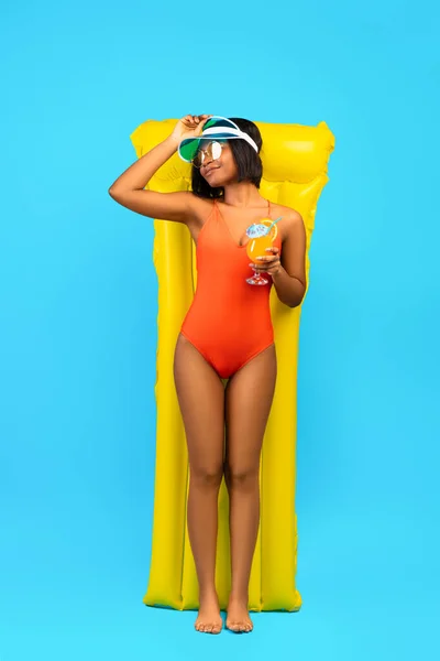 Full length of relaxed black lady in swimsuit lying on yellow inflatable lilo and drinking cocktail over blue background — Stock Photo, Image