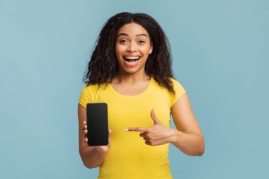 Mavi stüdyo arka planında boş ekranlı akıllı telefonu işaret eden neşeli siyahi bayan, mobil uygulama tasarımı için model