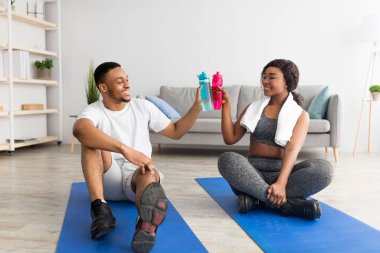 Formda siyahi erkek ve kız arkadaş spor şişeleriyle tezahürat yapıyor, yoga minderlerine oturuyor, antremandan sonra su içiyor.