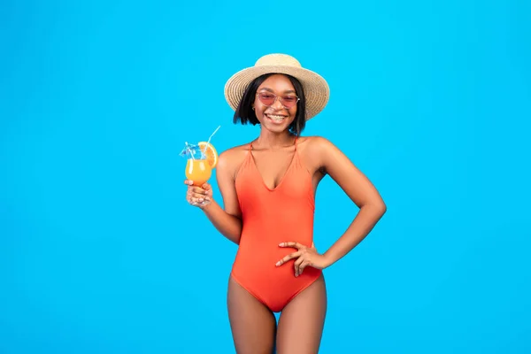 Aantrekkelijke zwarte dame in bikini en strohoed met glas koele cocktail op blauwe studio achtergrond — Stockfoto