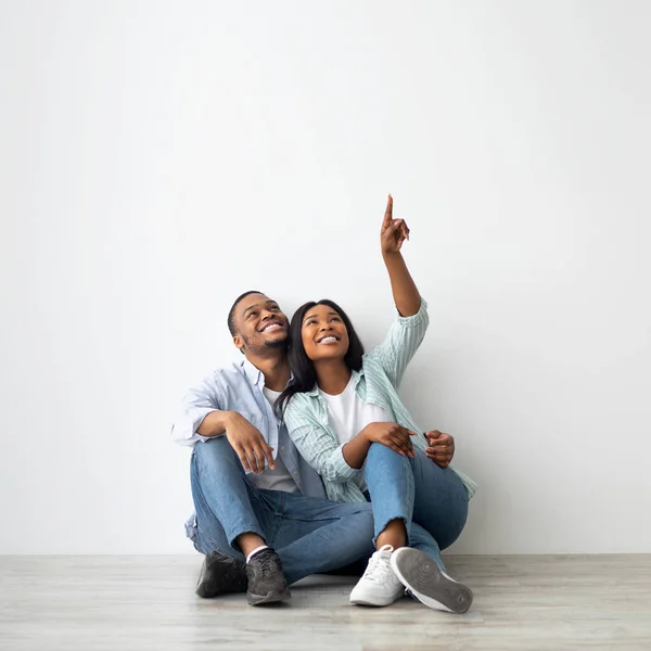 Anúncio para alugar apartamento. Felizes cônjuges afro-americanos sentados no chão na sala de estar sobre a parede branca — Fotografia de Stock