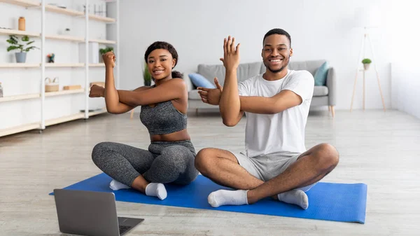 Онлайн-клас йоги. Молода чорна пара розтягує руки, сидячи в позі лотоса, тренуючись разом вдома, використовуючи ноутбук — стокове фото
