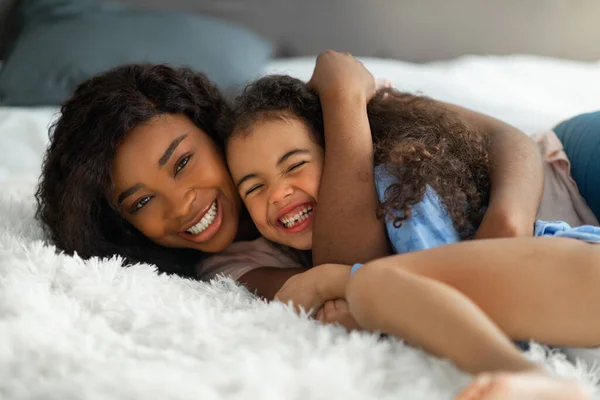 Die Liebe der Eltern. Glückliche junge schwarze Frau umarmt ihre kleine Tochter auf dem Bett, süßes Mädchen lacht und genießt die Umarmung — Stockfoto