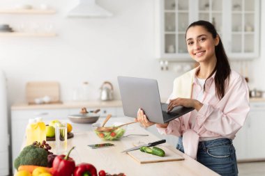 Kadın yemek pişiriyor ve dizüstü bilgisayar ve mutfak kullanıyor.