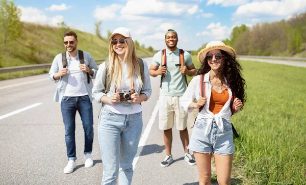 Grupo de jovens amigos diversificados com mochilas andando ao longo da estrada, viajando juntos fora — Fotografia de Stock