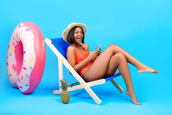 Belle femme noire se détendre dans une chaise longue, en utilisant un smartphone, discuter avec un ami en ligne sur la plage, fond bleu — Photo