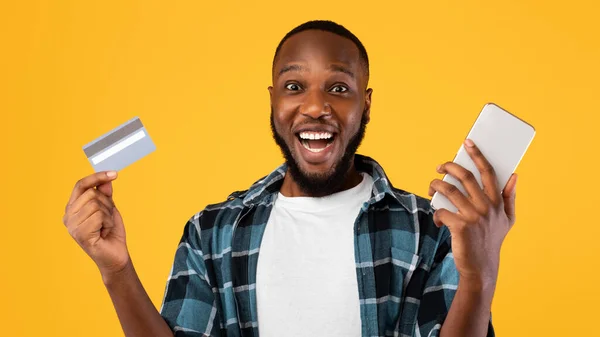 Podekscytowany czarny facet przy użyciu karty kredytowej i smartfona, żółte tło — Zdjęcie stockowe