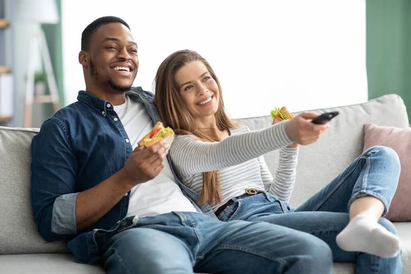 Χαρούμενο νεαρό διαφυλετικό ζευγάρι τρώει σνακ και βλέπει τηλεόραση στο σαλόνι — Φωτογραφία Αρχείου