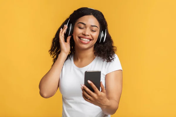 Les technologies modernes. Femme afro-américaine écouter de la musique dans les écouteurs, tenant smartphone sur fond jaune — Photo