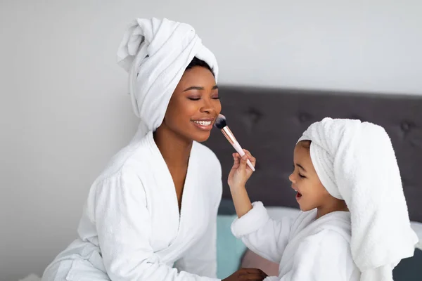 Bela mãe e filha negra em roupões de banho e toalhas aplicando cosméticos na cama em casa — Fotografia de Stock