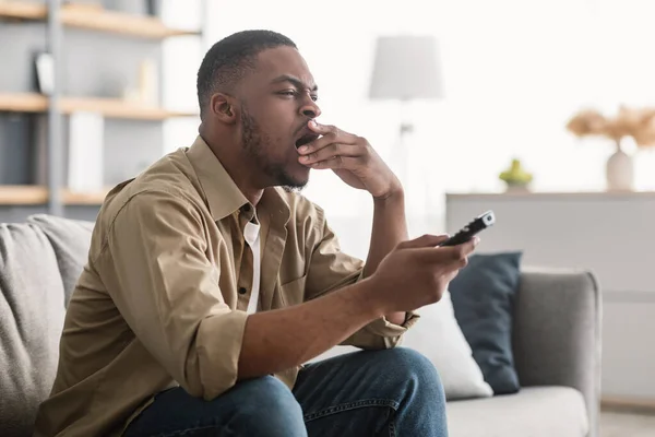 Скучный черный парень зевает, смотря телевизор, сидя на диване в помещении — стоковое фото