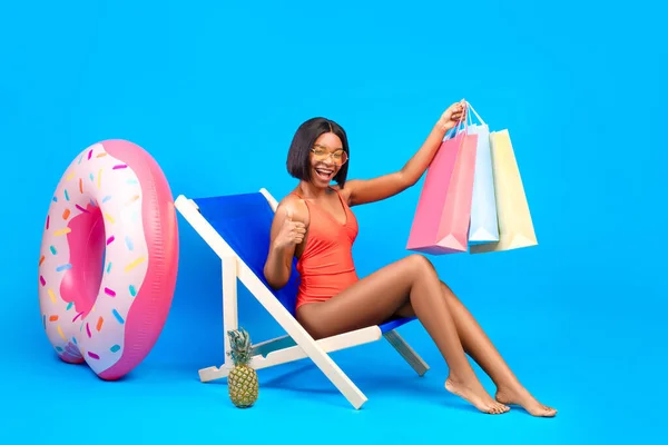 Venda de verão. Comprimento total da mulher negra sentada na cadeira lounge, segurando sacos de compras, mostrando o polegar, fundo azul — Fotografia de Stock