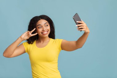 Heyecanlı Afrikalı Amerikalı kadın akıllı telefonuyla selfie çekiyor ve mavi stüdyo geçmişinde zafer jesti gösteriyor.