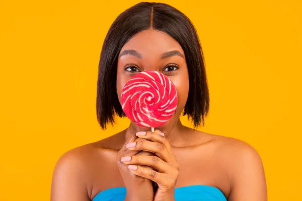 Lovely black woman hiding behind tasty striped lollipop on orange studio background — Stock Photo, Image