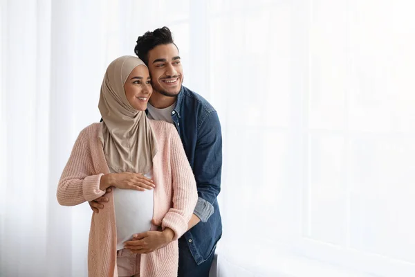 Portrait Of Happy Pregnant Islamic Lady And Her Arab Husband Hugging Near Window At Home — Stok Foto