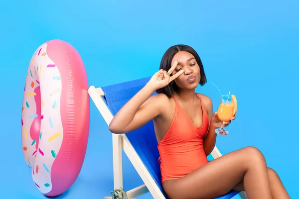 Flirty black lady with tropical cocktail relaxing in lounge chair, pursing her lips for kiss, making peace gesture