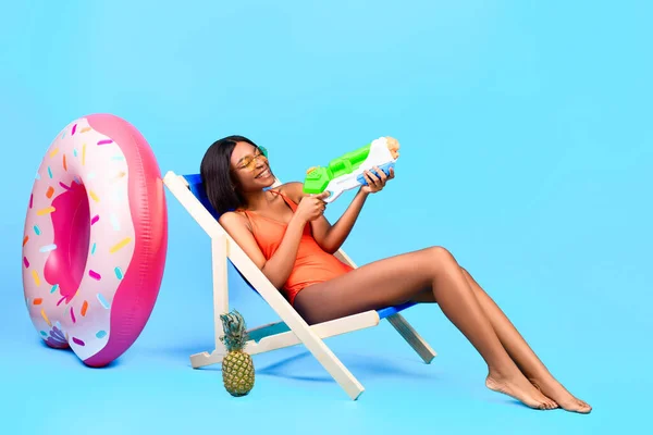 Amusant l'été. Belle femme noire assise dans une chaise longue avec pistolet à eau, jouant au jeu de plage sur fond bleu — Photo