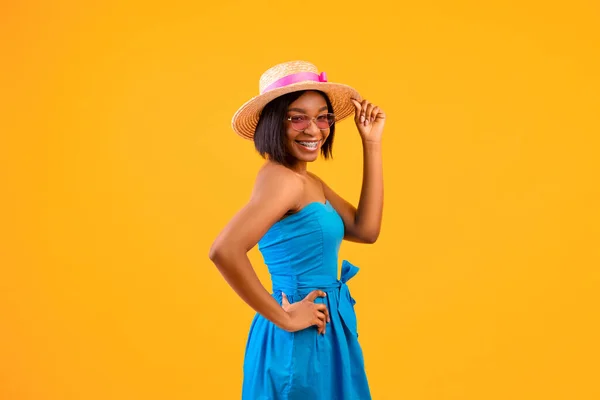 Şık mavi elbiseli genç siyahi bayan hasır şapkasına dokunuyor, turuncu arka planda sahile gidiyor. — Stok fotoğraf