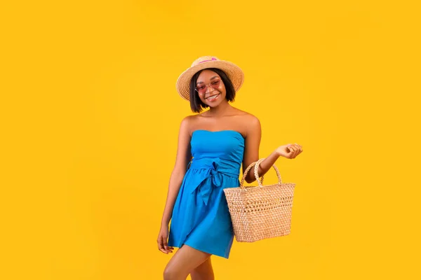 Portrait of young pretty black lady in straw hat and blue dress smiling, holding trendy beach bag over orange background — Stock Photo, Image