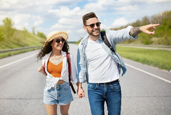 Glada unga par med ryggsäckar promenader längs vägen, pekar åt sidan, håller händerna, liftar tillsammans på sommaren — Stockfoto