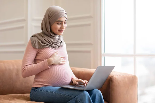 Embarazada musulmana mujer usando portátil sentado en sofá en casa — Foto de Stock