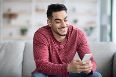 Gülümseyen Arap adam kanepede oturuyor, akıllı telefon kullanıyor.