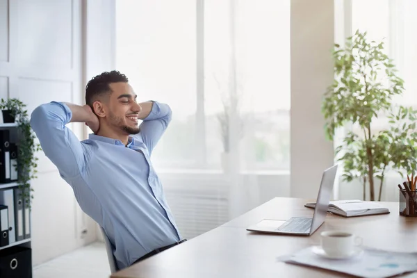 Σίγουρος Άραβας υπάλληλος της εταιρείας ακουμπά στην καρέκλα με τα χέρια πίσω από το κεφάλι, χαλαρώνοντας μετά την ολοκλήρωση της εργασίας στο χώρο εργασίας — Φωτογραφία Αρχείου