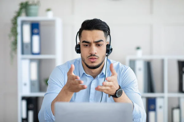 Hombre árabe joven preocupado en auriculares haciendo videollamada en línea con su socio de negocios, resolviendo problemas en la oficina —  Fotos de Stock