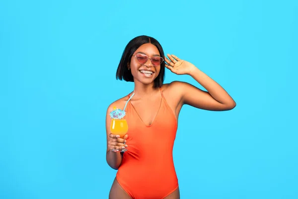 Summertime chill. Glamorous black lady in swimwear holding tropical fruit cocktail, smiling at camera, blue background — Stock Photo, Image