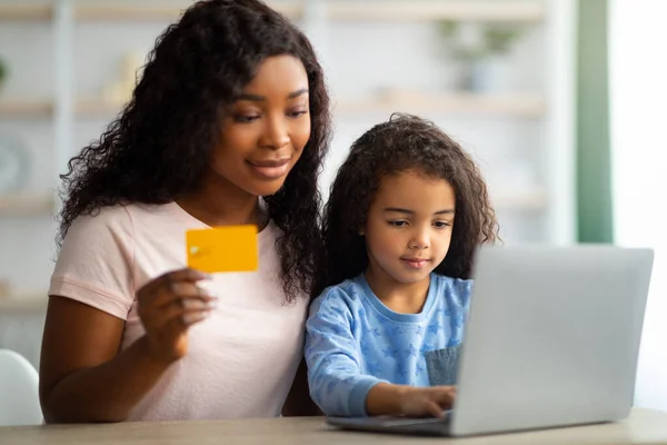 African American mother shopping online with credit card, making purchases on internet with her daughter from home