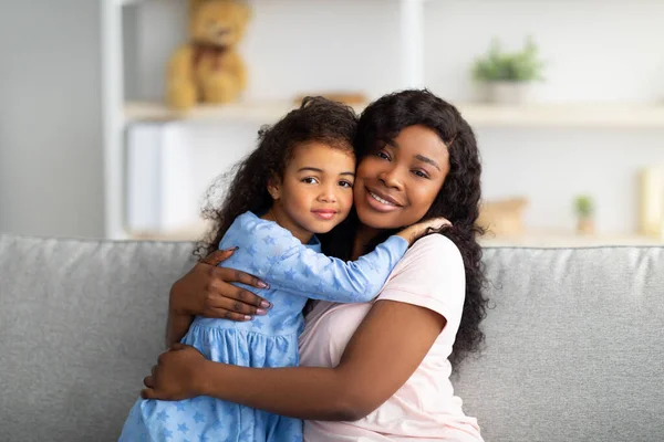 Liebevolles schwarzes Mädchen in hübschem Kleid, das ihre Mutter auf dem Sofa im Wohnzimmer umarmt. Starkes Konzept für Familienbeziehungen — Stockfoto