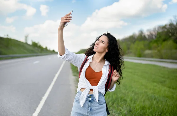 Mujer joven molesta caminando por la carretera, levantando la mano con el teléfono inteligente, buscando señal, sin conexión, al aire libre — Foto de Stock